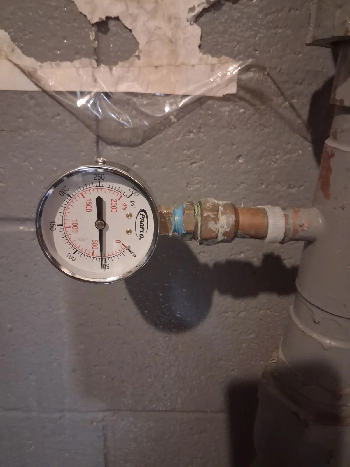 A round water-pressure gauge connected to utility-room plumbing against a block wall.